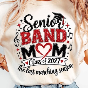 May include: A cream-colored t-shirt featuring a graphic with the text Senior BAND MOM, a graduation cap, musical notes, and a red heart. Below, a banner reads Class of 2027, followed by the text The last marching season in black script.