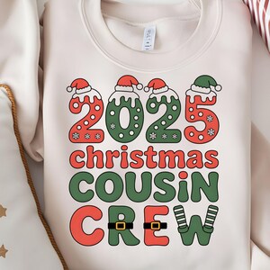 May include: Cream-colored sweatshirt with a festive graphic. The design features the numbers "2025" in red and green, topped with Santa hats, and the words "Christmas Cousin Crew". A holiday-themed apparel item.