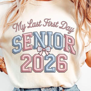 Peut inclure: T-shirt crème avec l'inscription "My Last First Day SENIOR 2026" en rose et bleu. Le mot "SENIOR" est dans un style de lettres en blocs avec un nœud au centre. L'année "2026" est en rose.