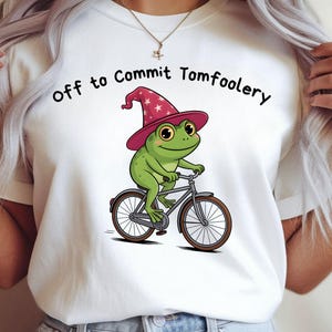 May include: White t-shirt with a cartoon frog on a bicycle, wearing a pink wizard hat with white stars. The frog is green with yellow eyes. The text "Off to Commit Tomfoolery" is printed above the frog.