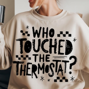 May include: A beige sweatshirt with the text "WHO TOUCHED THE THERMOSTAT?" in black. The text is in a bold, playful font with decorative elements. A black mug is held in the person's hand.