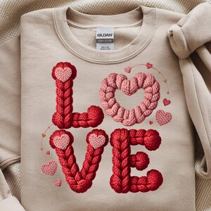 Peut inclure: Un sweat-shirt beige avec le mot "LOVE" en tricot. Les lettres sont rouges et roses, avec des cœurs. Le sweat-shirt est plié sur une couverture beige.