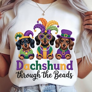 May include: White t-shirt featuring three dachshunds dressed in Mardi Gras attire. Each dog wears a different hat and sweater, with the text "Dachshund through the Beads" in colorful lettering. The dogs are in a row, each with a unique expression.