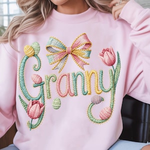 Peut inclure: Un sweat-shirt rose clair avec le mot "Granny" brodé en fil coloré, orné d'un nœud, d'œufs de Pâques et de tulipes. Le motif présente des couleurs pastel et un thème printanier.
