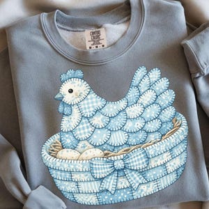 Peut inclure: Un sweat-shirt bleu clair avec une illustration détaillée d'une poule dans un panier avec des œufs. La poule et le panier sont décorés de motifs patchwork bleus et blancs. Le sweat-shirt a un col rond et des manches longues.