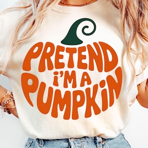 Peut inclure: T-shirt crème avec l'inscription orange "PRETEND I'M A PUMPKIN" formant un motif de citrouille, surmontée d'une tige verte. Vêtement décontracté sur le thème d'Halloween et de l'automne.