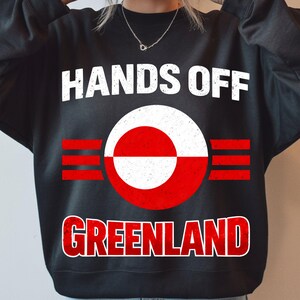 Peut inclure: Sweat-shirt noir avec l'inscription "HANDS OFF GREENLAND" en blanc et rouge. Le motif comprend un cercle rouge et blanc avec des rayures rouges horizontales, représentant le drapeau du Groenland.