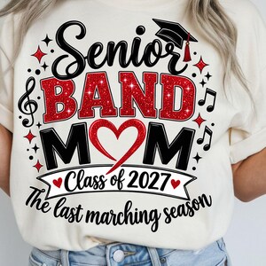 May include: A cream-colored t-shirt featuring a graphic with the text Senior BAND MOM, Class of 2027, and The last marching season. The design includes a black graduation cap, red glitter lettering, a red heart, and black musical notes.