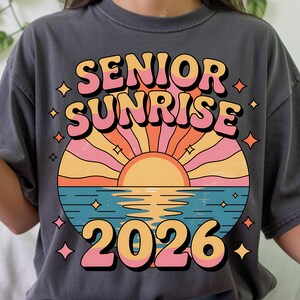 May include: Dark gray t-shirt with a colorful graphic. The graphic features the words "SENIOR SUNRISE 2026" in a retro font, with a sunset design in shades of orange, pink, and yellow over blue water. The shirt has a relaxed fit.