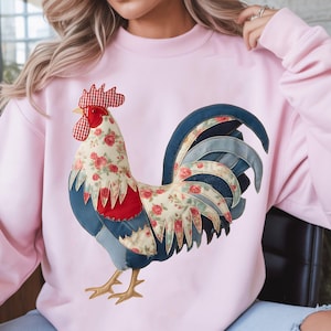 Peut inclure: Un sweat-shirt rose clair avec un motif de coq. Le coq a une crête rouge, un corps à motifs floraux et une queue à motifs bleus et floraux. Le sweat-shirt est de couleur rose clair uni.
