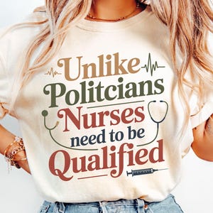 Puede incluir: Camiseta color crema con el texto "Unlike Politicians Nurses need to be Qualified" en letras marrones, verdes y rojas. El diseño incluye un estetoscopio, una línea de latidos y una jeringa.