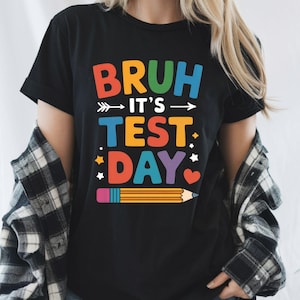 Peut inclure: T-shirt noir avec l'inscription colorée "BRUH IT'S TEST DAY". Le motif comprend une flèche, des étoiles, un cœur et un crayon. Un vêtement décontracté.