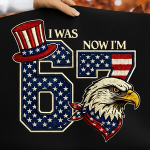 Peut inclure: Graphique noir avec le texte "I WAS 67 NOW I'M", présentant un design patriotique. Il comprend un chapeau haut de forme rouge, blanc et bleu, le chiffre 67 rempli d'étoiles et un aigle portant un bandana.