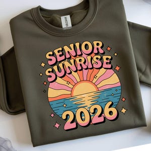 May include: Olive green sweatshirt with a retro graphic that reads "SENIOR SUNRISE 2026". The design features a colorful sunset over water with a retro font. The sweatshirt is a Gildan brand.