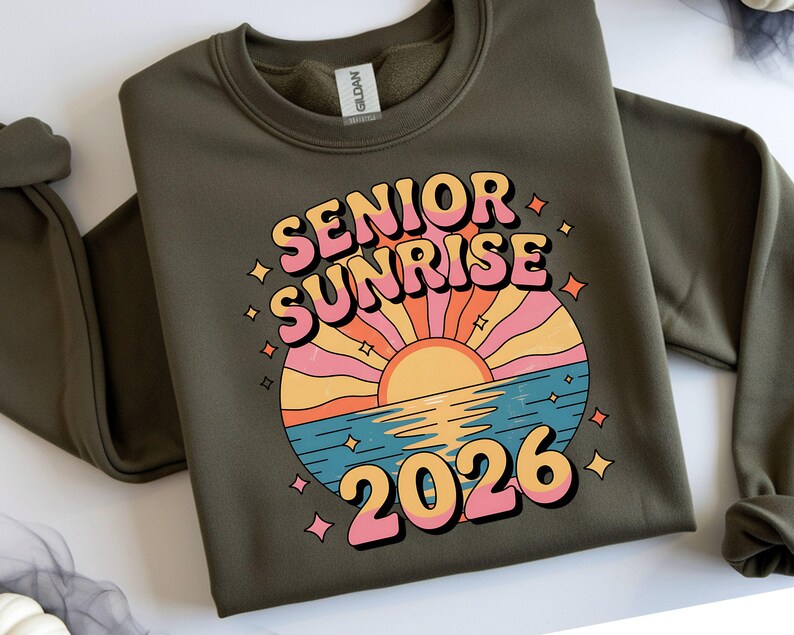 May include: Olive green sweatshirt with a retro graphic that reads "SENIOR SUNRISE 2026". The design features a colorful sunset over water with a retro font. The sweatshirt is a Gildan brand.