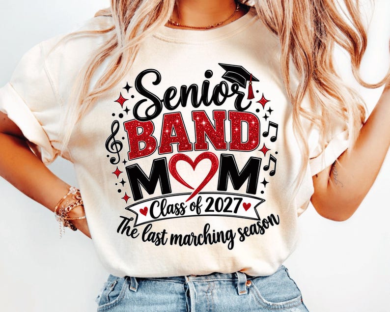 May include: A cream-colored t-shirt featuring a graphic with the text Senior BAND MOM, a graduation cap, musical notes, and a red heart. Below, a banner reads Class of 2027, followed by the text The last marching season in black script.