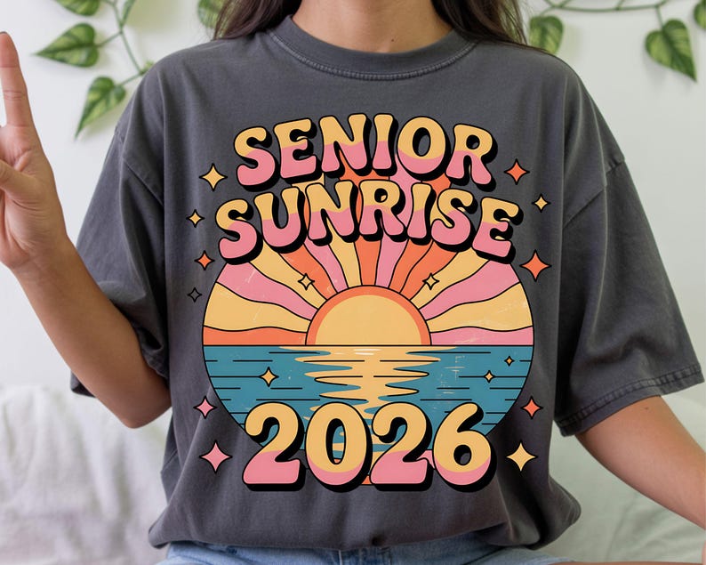 May include: Dark gray t-shirt with a colorful graphic. The graphic features the words "SENIOR SUNRISE 2026" in a retro font, with a sunset design in shades of orange, pink, and yellow over blue water. The shirt has a relaxed fit.