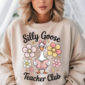 Może przedstawiać: Beżowa bluza z napisem "Silly Goose Teacher Club" i kresk&oacute;wkową gęsią w okularach i kokardzie, trzymającą książkę. Projekt zawiera r&oacute;żowe, ż&oacute;łte i białe kwiaty oraz małe złote serca.