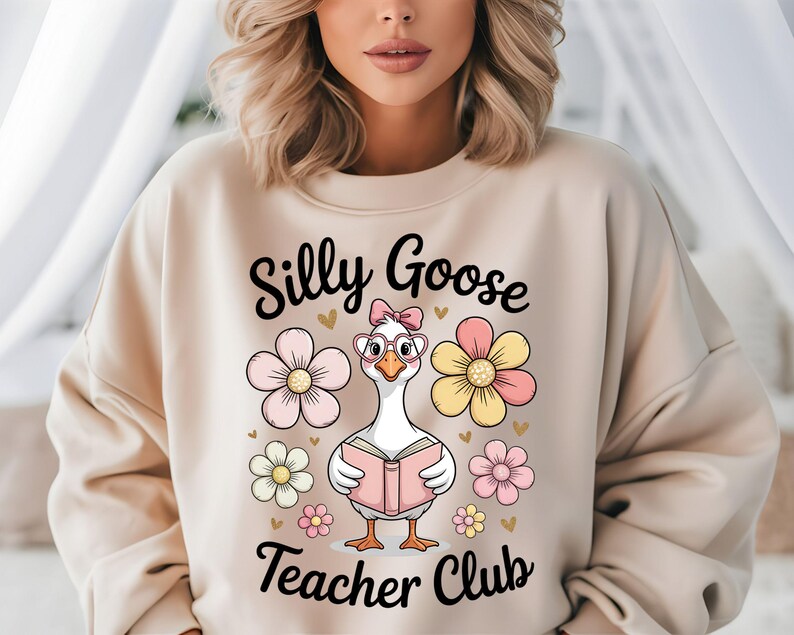 Może przedstawiać: Beżowa bluza z napisem "Silly Goose Teacher Club" i kresk&oacute;wkową gęsią w okularach i kokardzie, trzymającą książkę. Projekt zawiera r&oacute;żowe, ż&oacute;łte i białe kwiaty oraz małe złote serca.