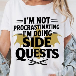 May include: White t-shirt with black text that reads "I'm not procrastinating I'm doing side quests" with a gold dragon design and two crossed swords.