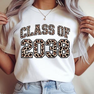 May include: White t-shirt with the text "CLASS OF 2038" in a leopard print design. The text is outlined in black. The shirt is a crew neck style.