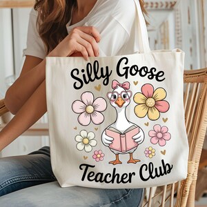 Może przedstawiać: Kremowa torba z napisem "Silly Goose Teacher Club". Torba przedstawia kresk&oacute;wkową gęś w okularach i kokardzie, trzymającą książkę, otoczoną r&oacute;żowymi i ż&oacute;łtymi kwiatami. Zabawny i fantazyjny design.