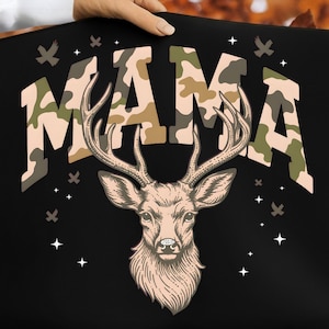 Peut inclure: Sac fourre-tout noir avec le mot "MAMA" en imprimé camouflage, avec une illustration détaillée d'une tête de cerf avec de grands bois. De petits graphiques d'oiseaux et d'étoiles entourent le texte et le cerf.