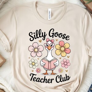 Może przedstawiać: Beżowa koszulka z napisem "Silly Goose Teacher Club" i kresk&oacute;wkową gęsią w okularach i z książką, otoczoną r&oacute;żowymi i ż&oacute;łtymi kwiatami. Koszulka jest wykonana z miękkiego materiału.