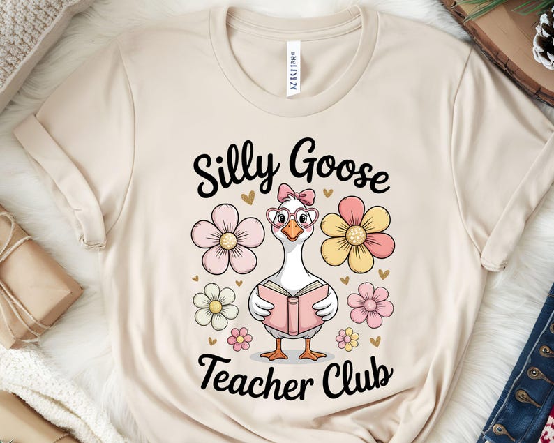 Może przedstawiać: Beżowa koszulka z napisem "Silly Goose Teacher Club" i kresk&oacute;wkową gęsią w okularach i z książką, otoczoną r&oacute;żowymi i ż&oacute;łtymi kwiatami. Koszulka jest wykonana z miękkiego materiału.