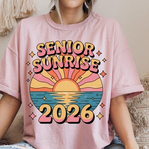 May include: A light pink t-shirt featuring the words "SENIOR SUNRISE 2026" in a retro font. The design includes a colorful sunset over water with diamond accents, creating a vintage aesthetic.