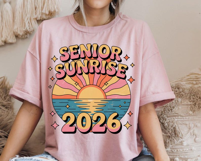 May include: A light pink t-shirt featuring the words "SENIOR SUNRISE 2026" in a retro font. The design includes a colorful sunset over water with diamond accents, creating a vintage aesthetic.