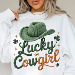 Peut inclure: Sweat-shirt blanc avec un chapeau de cowboy vert tricoté et l'inscription "Lucky Cowgirl" en vert et orange. Le motif comprend des trèfles et des étoiles, idéal pour la Saint-Patrick ou un thème western.