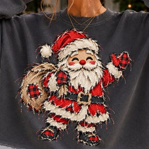 May include: A charcoal gray crewneck sweatshirt featuring a large, frayed-edge applique of Santa Claus. Santa wears a red suit with black and red buffalo plaid accents on the mittens, boots, and toy sack, finished with a black belt and gold buckle.