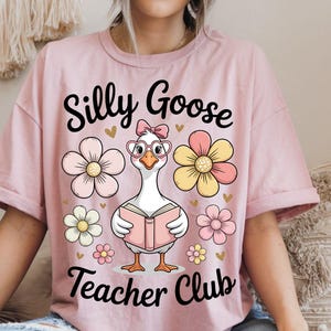 Może przedstawiać: R&oacute;żowa koszulka z napisem "Silly Goose Teacher Club". Na koszulce znajduje się rysunkowa gęś w okularach i kokardzie, trzymająca książkę, otoczona kwiatami.