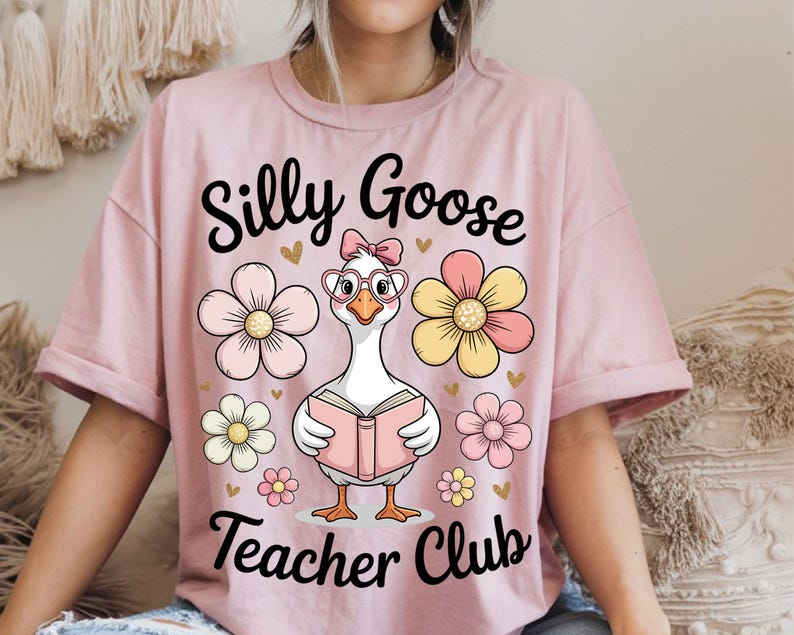 Może przedstawiać: R&oacute;żowa koszulka z napisem "Silly Goose Teacher Club". Na koszulce znajduje się rysunkowa gęś w okularach i kokardzie, trzymająca książkę, otoczona kwiatami.