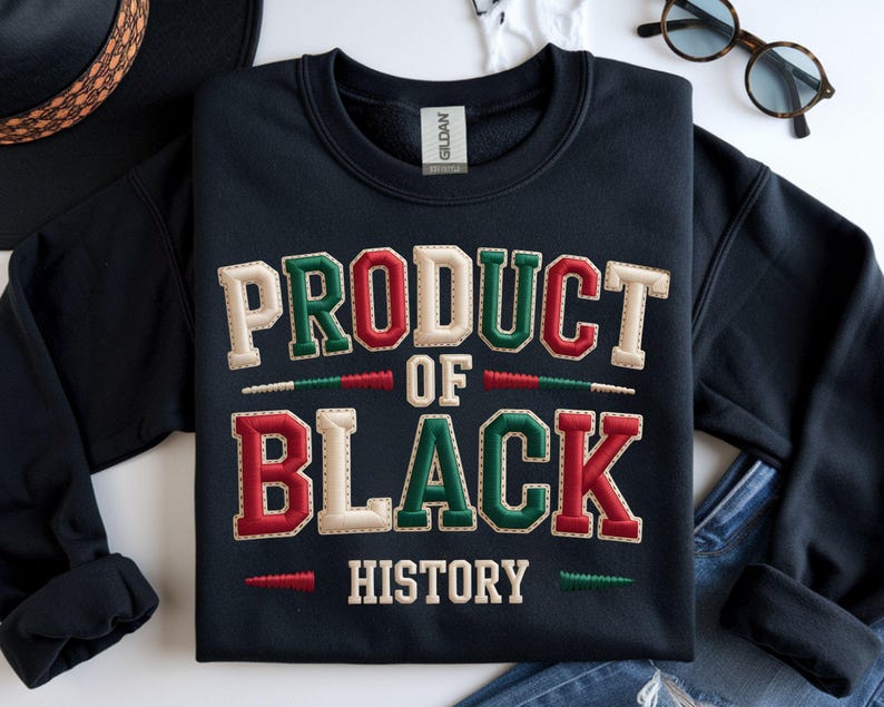 May include: Black sweatshirt featuring the text "PRODUCT OF BLACK HISTORY" in red, green, and cream-coloured lettering. The sweatshirt is folded, with a hat and glasses in the background.