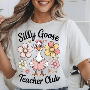 Może przedstawiać: Biała koszulka z napisem "Silly Goose Teacher Club". Koszulka przedstawia kresk&oacute;wkową gęś w okularach i kokardzie, trzymającą książkę, otoczoną kwiatami i sercami.