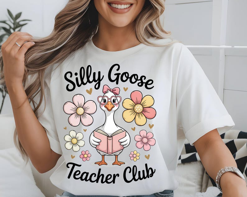 Może przedstawiać: Biała koszulka z napisem "Silly Goose Teacher Club". Koszulka przedstawia kresk&oacute;wkową gęś w okularach i kokardzie, trzymającą książkę, otoczoną kwiatami i sercami.