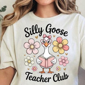 Może przedstawiać: Kremowa koszulka z wizerunkiem kresk&oacute;wki gęsi w okularach i kokardzie, trzymającej r&oacute;żową książkę. Tekst "Silly Goose Teacher Club" znajduje się nad i pod gęsią, otoczony kwiatami.