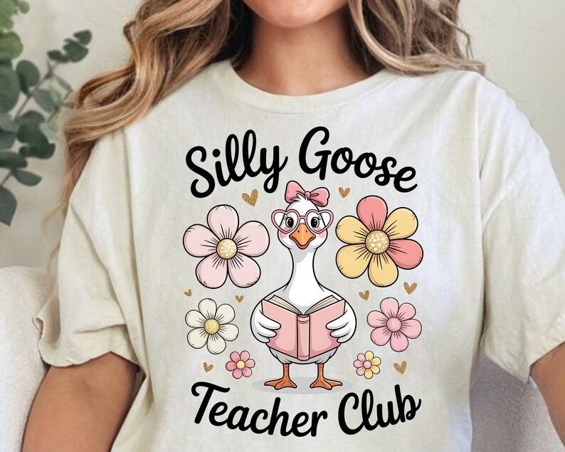 Może przedstawiać: Kremowa koszulka z wizerunkiem kresk&oacute;wki gęsi w okularach i kokardzie, trzymającej r&oacute;żową książkę. Tekst "Silly Goose Teacher Club" znajduje się nad i pod gęsią, otoczony kwiatami.