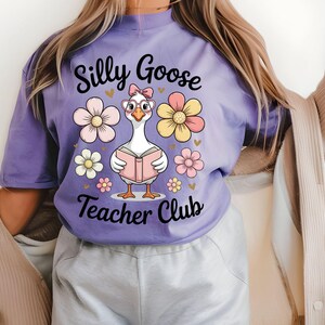 Może przedstawiać: Liliowa koszulka z kresk&oacute;wkową gęsią w okularach i kokardą, czytającą książkę. Tekst "Silly Goose Teacher Club" znajduje się nad i pod gęsią, z kwiatowymi i sercowymi akcentami.