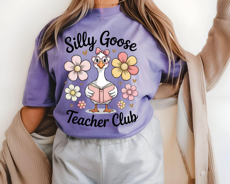 Może przedstawiać: Liliowa koszulka z kresk&oacute;wkową gęsią w okularach i kokardą, czytającą książkę. Tekst "Silly Goose Teacher Club" znajduje się nad i pod gęsią, z kwiatowymi i sercowymi akcentami.