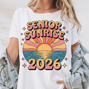 May include: White t-shirt featuring a retro design with the words "SENIOR SUNRISE" in a layered, colorful font. Below the text is a sunset scene over water, with the year "2026" in a matching font. The design includes star accents.