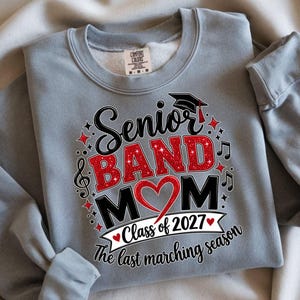 May include: A light gray crewneck sweatshirt featuring a graphic print that reads Senior BAND MOM with a red glitter heart, a graduation cap, musical notes, and the text Class of 2027 The last marching season in black and red lettering.