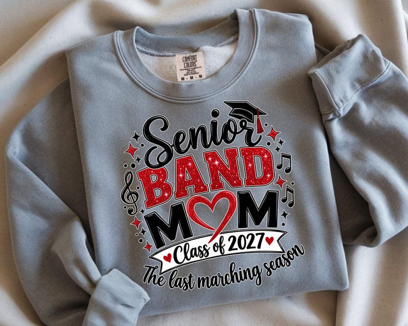 May include: A light gray crewneck sweatshirt featuring a graphic print that reads Senior BAND MOM with a red glitter heart, a graduation cap, musical notes, and the text Class of 2027 The last marching season in black and red lettering.