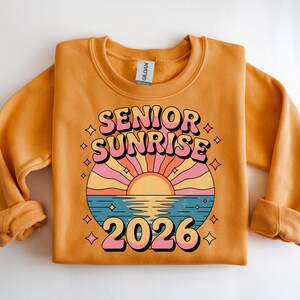 May include: Mustard-colored sweatshirt with the words "SENIOR SUNRISE 2026" in a retro font. The design features a sunset over water with pink, orange, and blue hues. The sweatshirt is made of a soft material.
