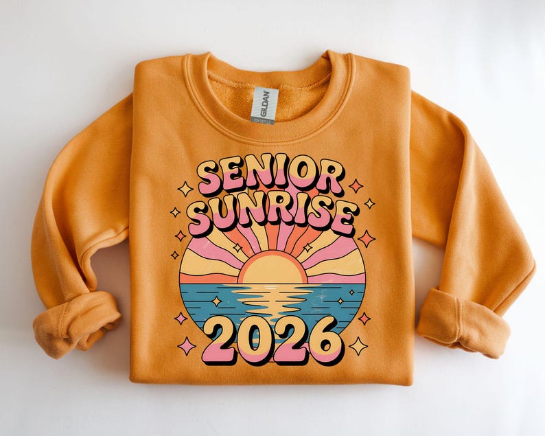 May include: Mustard-colored sweatshirt with the words "SENIOR SUNRISE 2026" in a retro font. The design features a sunset over water with pink, orange, and blue hues. The sweatshirt is made of a soft material.