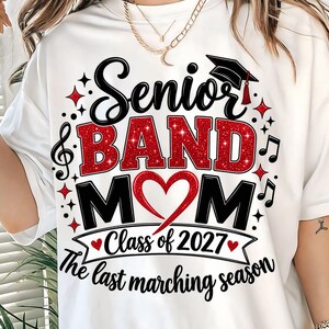 May include: A white t-shirt featuring a graphic design with the text Senior BAND MOM, Class of 2027, and The last marching season. The design includes a red glitter heart, a black graduation cap, musical notes, and red star accents.