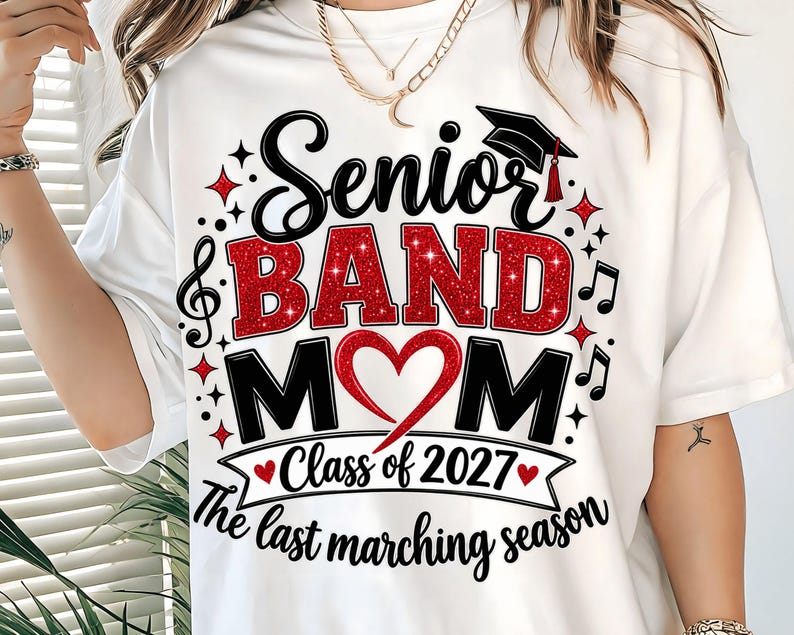 May include: A white t-shirt featuring a graphic design with the text Senior BAND MOM, Class of 2027, and The last marching season. The design includes a red glitter heart, a black graduation cap, musical notes, and red star accents.