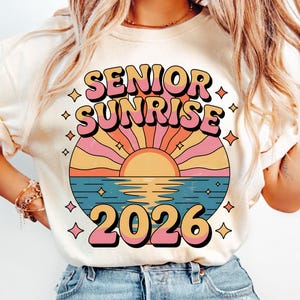 May include: Cream-colored t-shirt featuring a retro design with the words "SENIOR SUNRISE 2026" in pink and orange. The graphic includes a stylized sunset over water with a sun, rays, and stars.